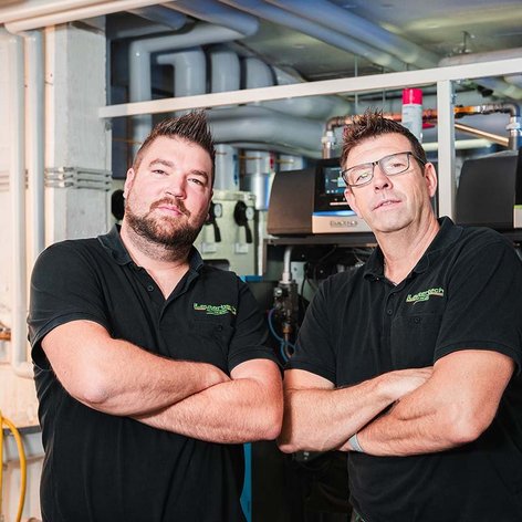 Zwei Männer in schwarzen T-Shirts stehen mit verschränkten Armen vor einer technischen Anlage in einem modernen Raum.