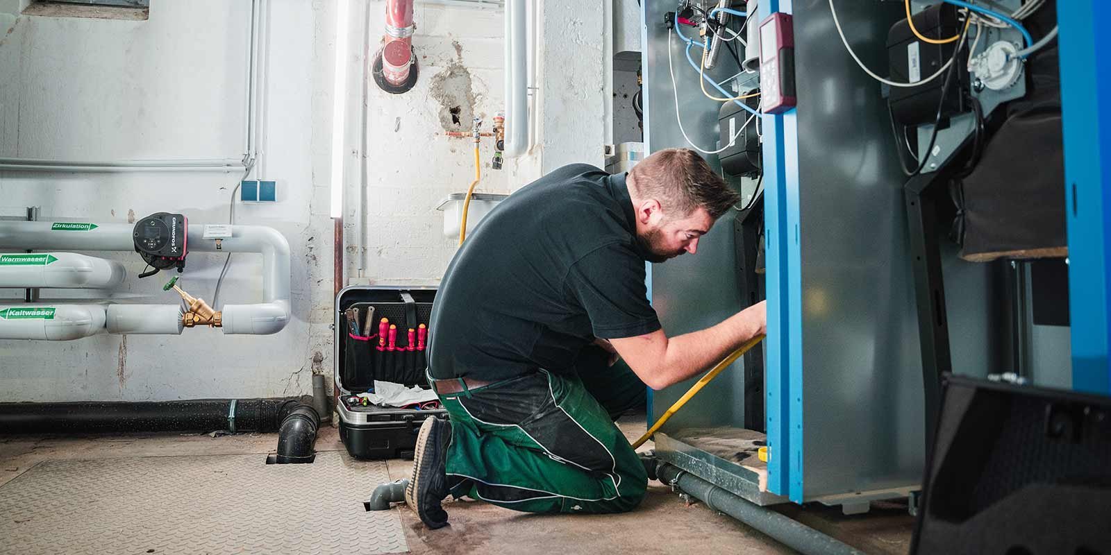 Ein Techniker kniet in einem Raum und arbeitet an einer blauen Maschine, umgeben von Rohren und Werkzeugen.