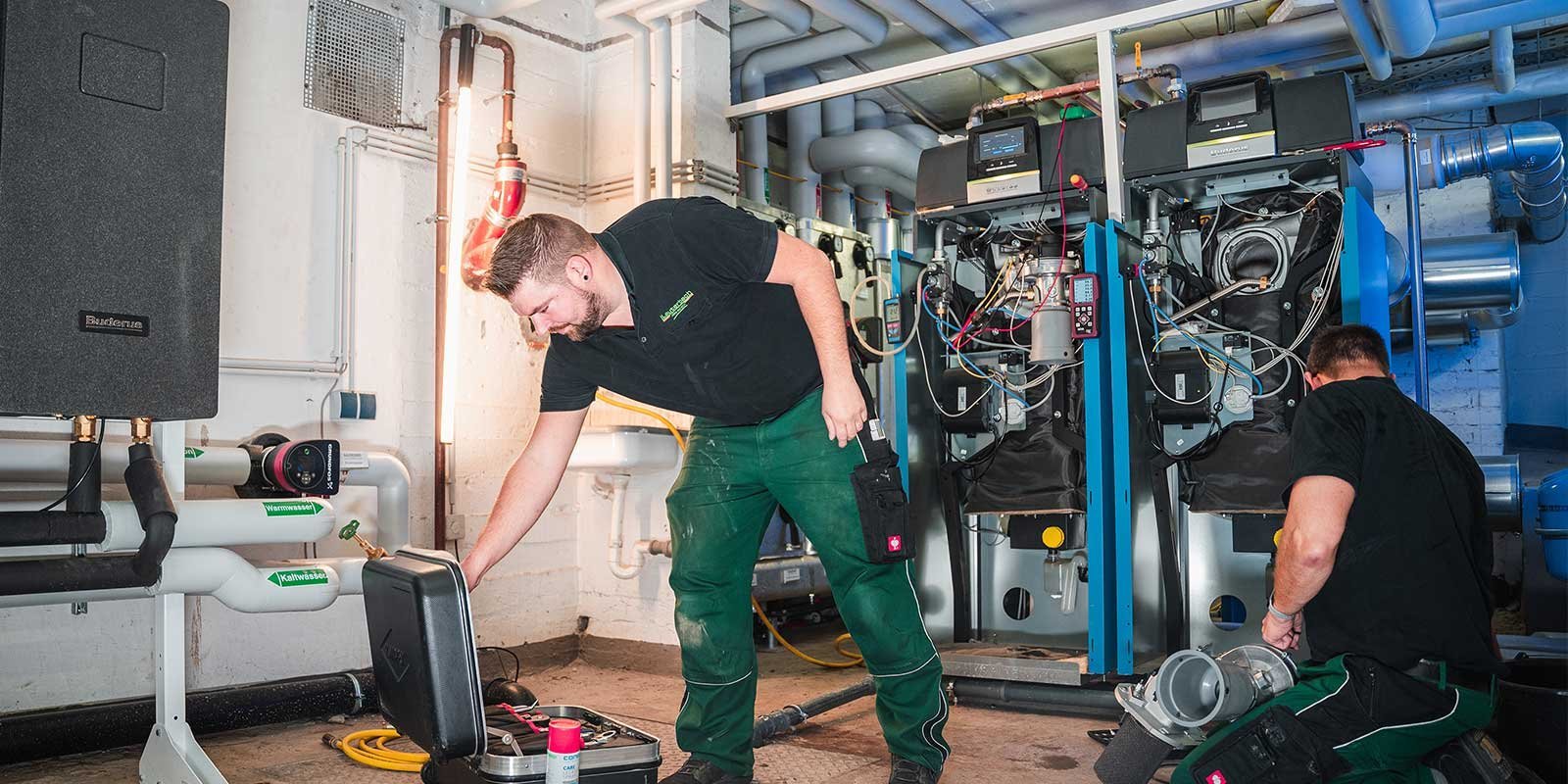Ein Techniker arbeitet konzentriert an einer Heizungsanlage, während ein Kollege im Hintergrund eine andere Maschine überprüft.